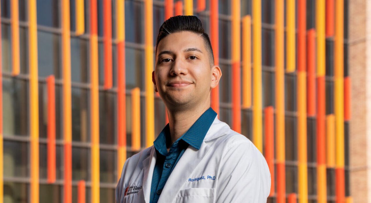 A man in a white lab coat smiles in front of an orange and yellow building.