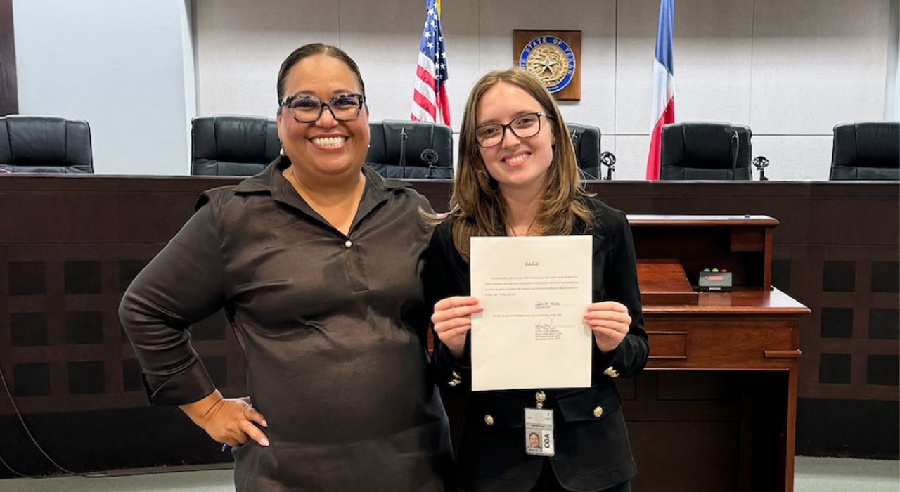 Two women stand in a court room smiling towards a camera. One is holding a piece of paper and the other stands beside.