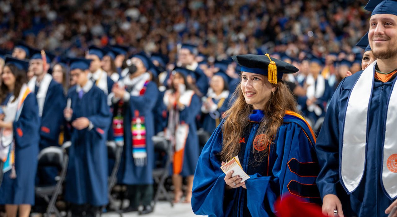 A woman in blue regalia to signify the type of degree she is earning stand amongst a crowd of people during a ceremony.