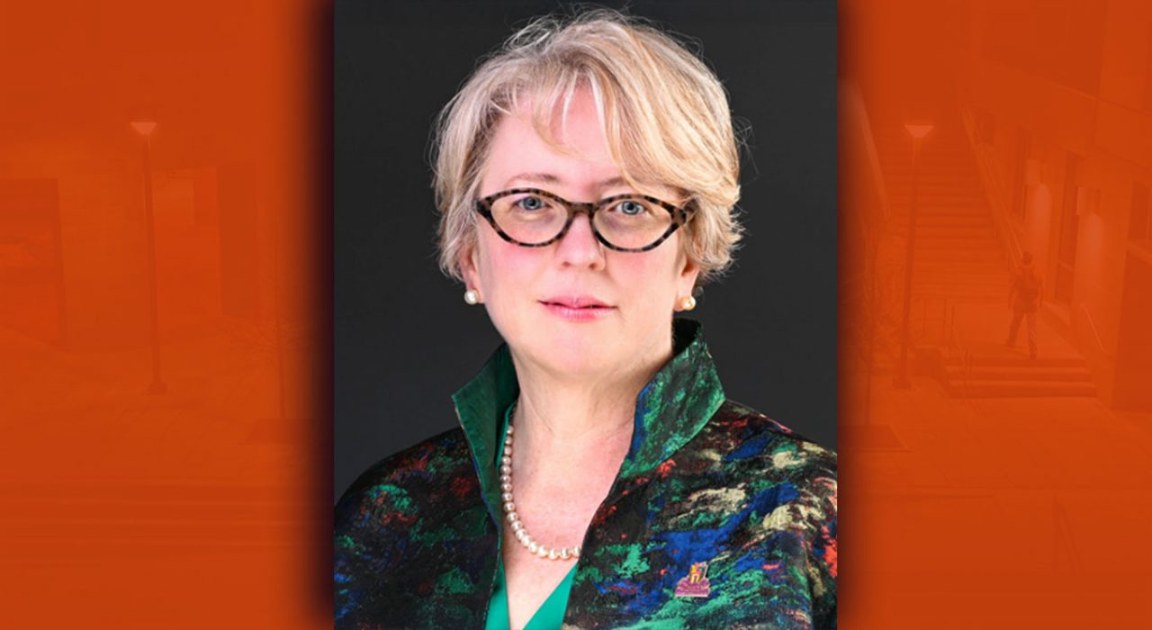 Portrait of a woman with short blonde hair, black glasses and a jacket with various colored splattered designs.