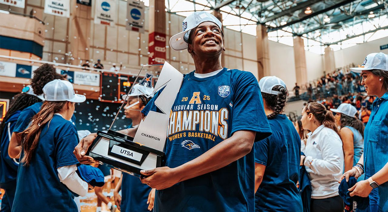 A female basketball player looks off to the side and carries a trophy as others celebrate behind her.