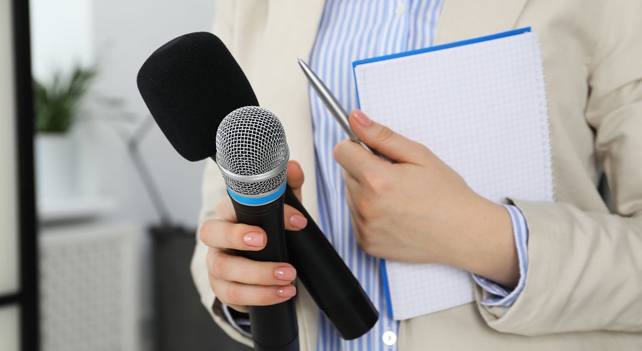 A closeup of a woman holding two microphones, a journal with a pen.