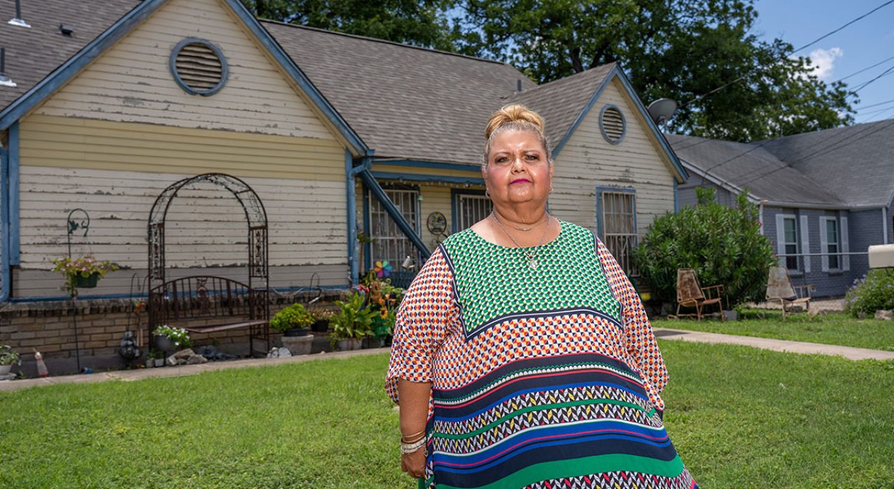 A woman in a flowy blouses stands at the forground in front of her house as the sun beams above her.