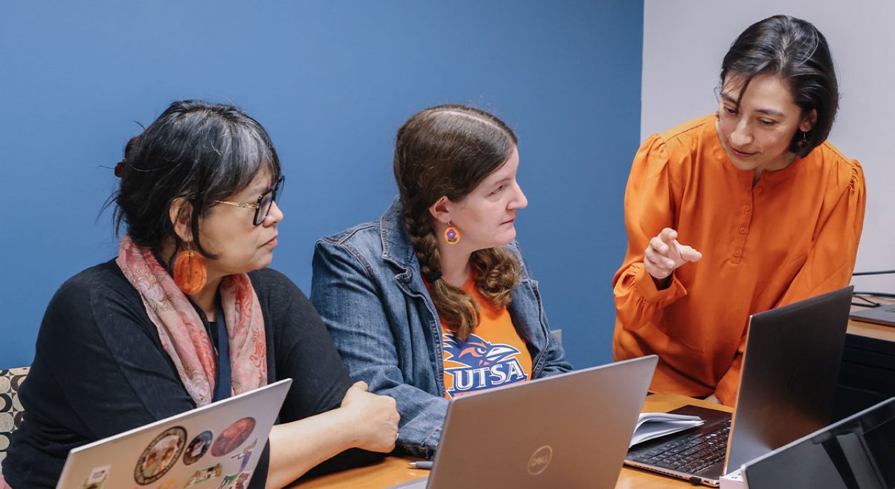 A woman in an orange blouse leans over a table as she talks to two other women on their laptops.
