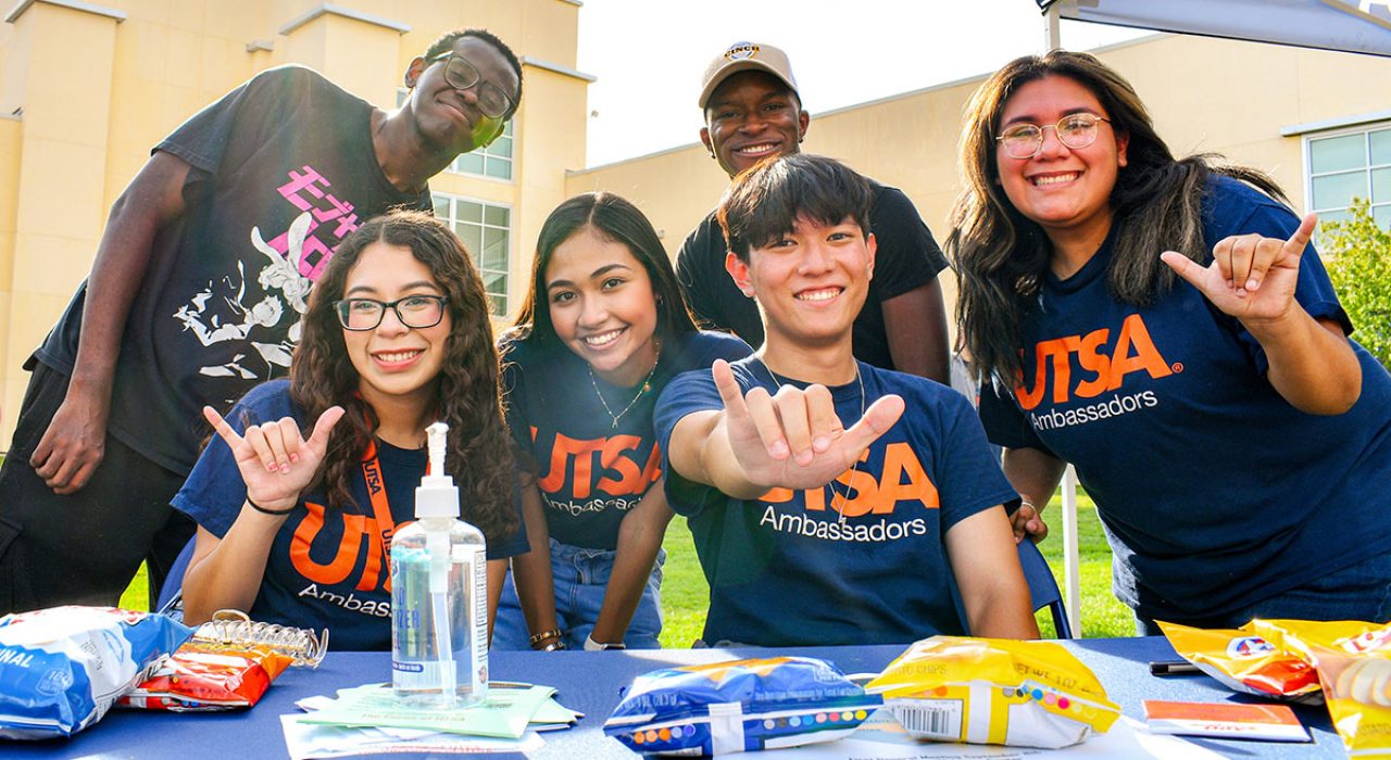Six UTSA student ambassadors are stationed behind a table with snacks and flyers