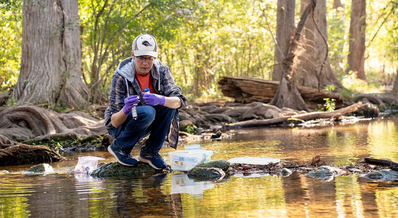 Ronny Saunders perches on a rock in the waters of Cibolo Creek while holding a vial for water testing