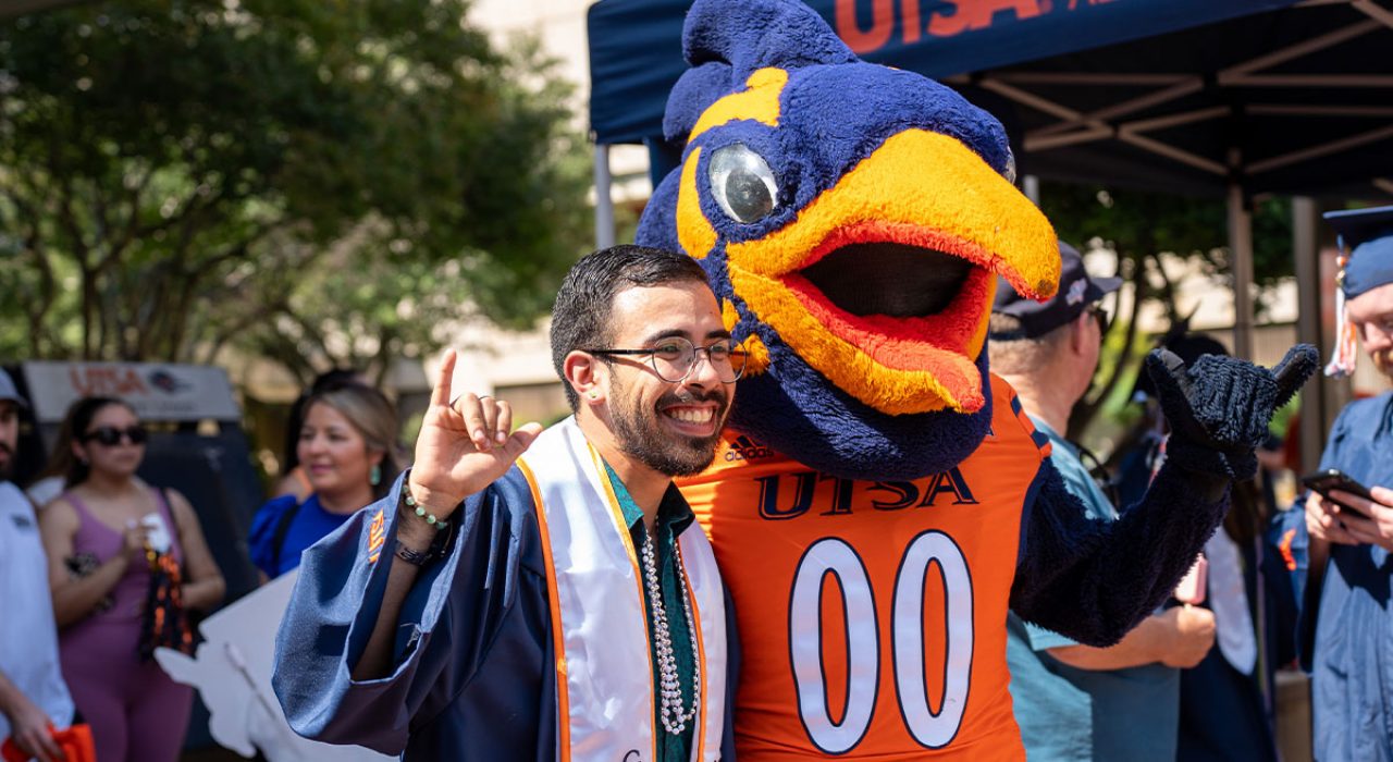 A mascot suit, which looks like a roadrunner stands with a man dressed in commencement regalia.