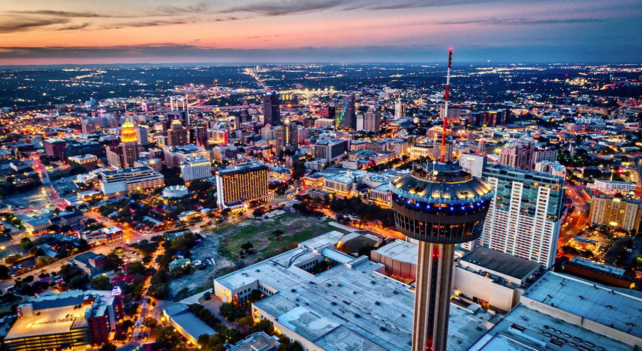 A bird's eye view of a city's downtown. The dozens of buildings are lit up and illuminated.