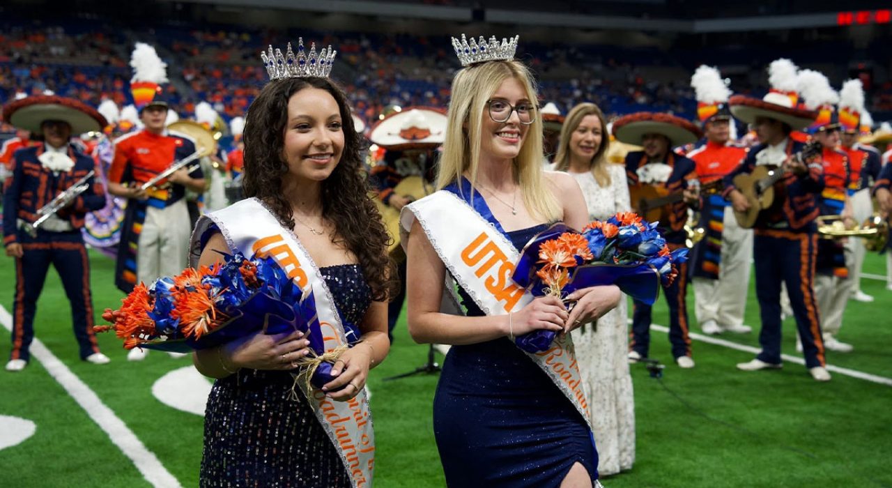 Two young women in formal gowns wear crowns and hold bouquets of flowers on a football field.
