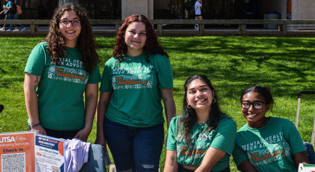 Four young woman in green shirts that say "Wellbeing" sit at a table outside.